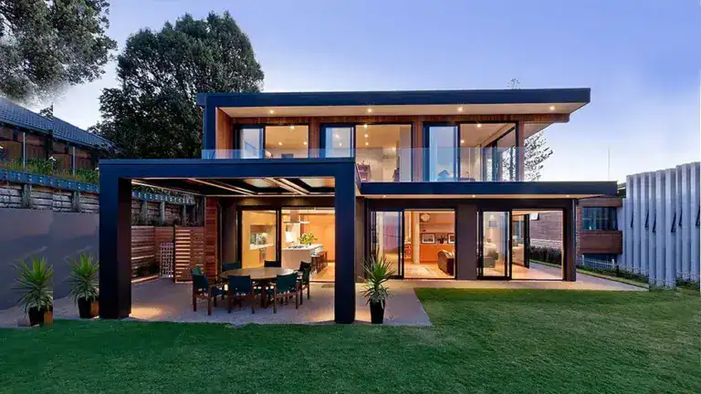 Modern two-story house with large windows, outdoor dining area, and lawn, lit up at dusk.