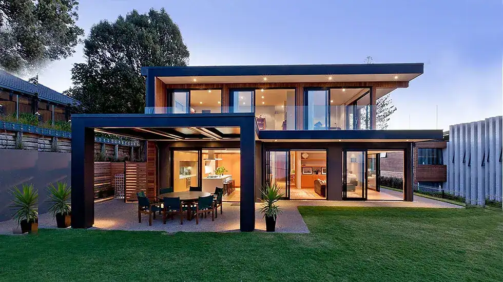 Modern two-story house with large windows, outdoor dining area, and lawn, lit up at dusk.