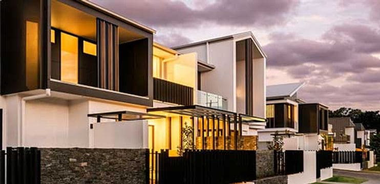Modern two-story houses with large windows, glowing interiors, and an overcast sky at dusk.