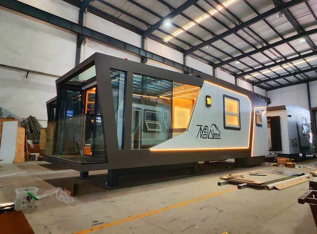A modern modular home with large windows and lit trim sits inside an industrial warehouse.