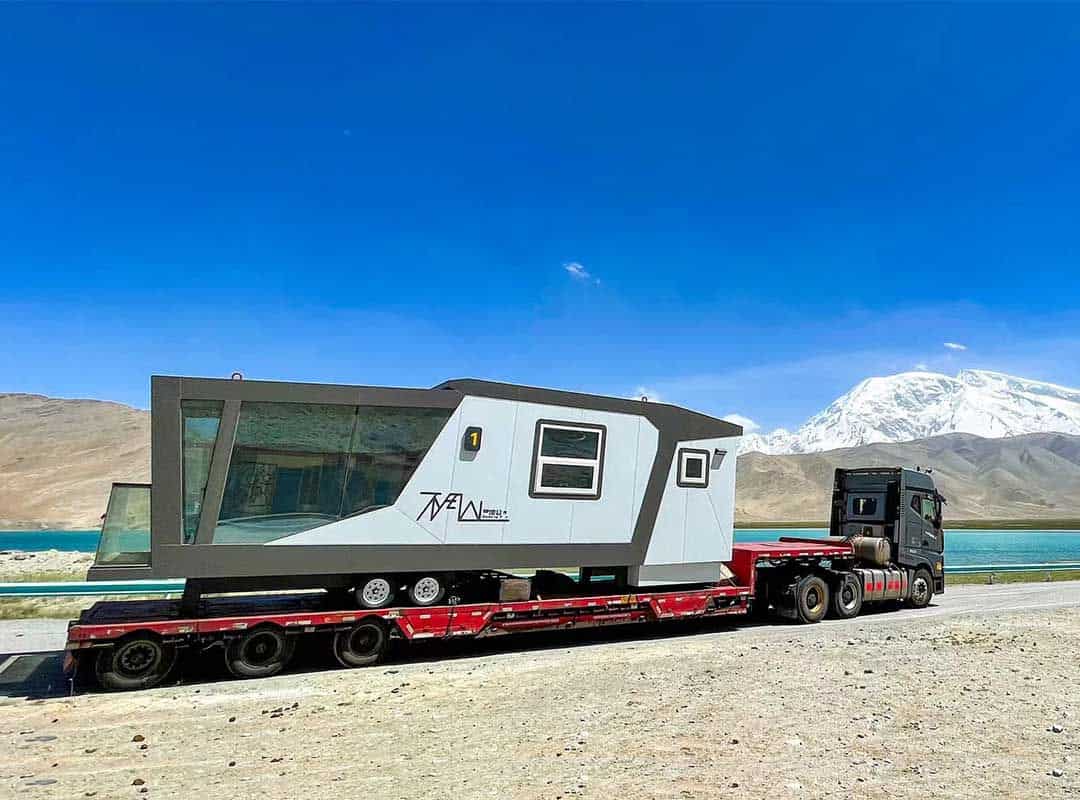 An angular mobile home rides on a flatbed truck by a lake, with mountains in the background.