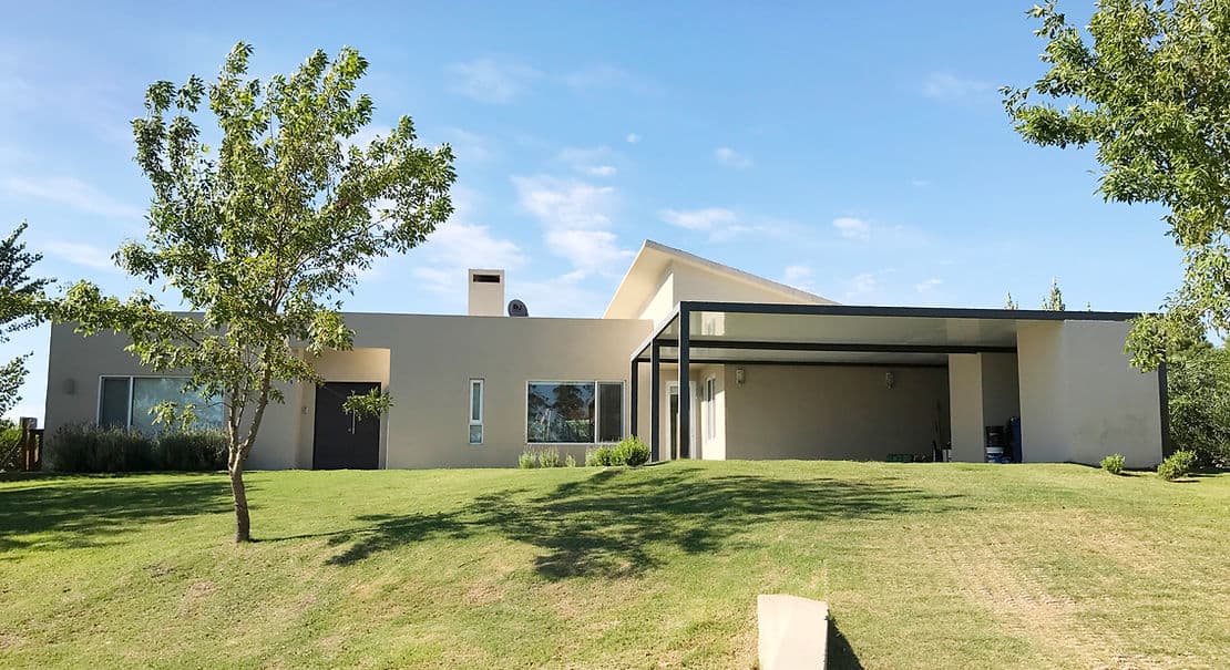 Modern one-story house with flat roof, large windows, and carport on grassy lawn with trees under a blue sky.