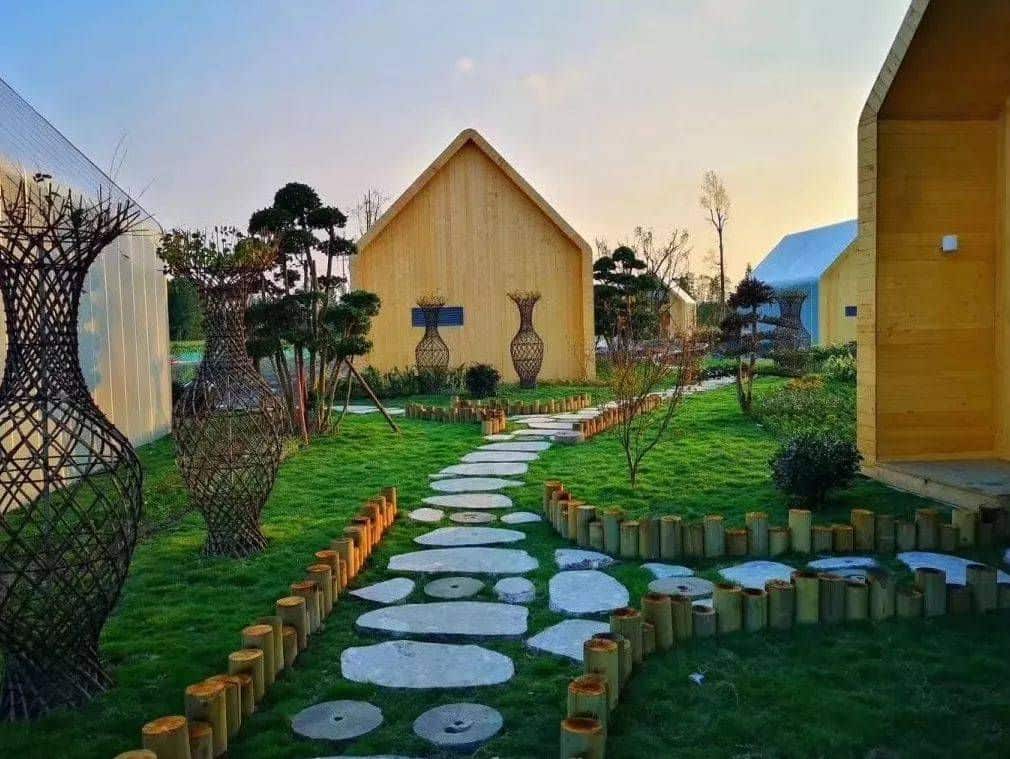 A stone path winds through a garden with sculptures and wooden cabins beneath a clear sky.