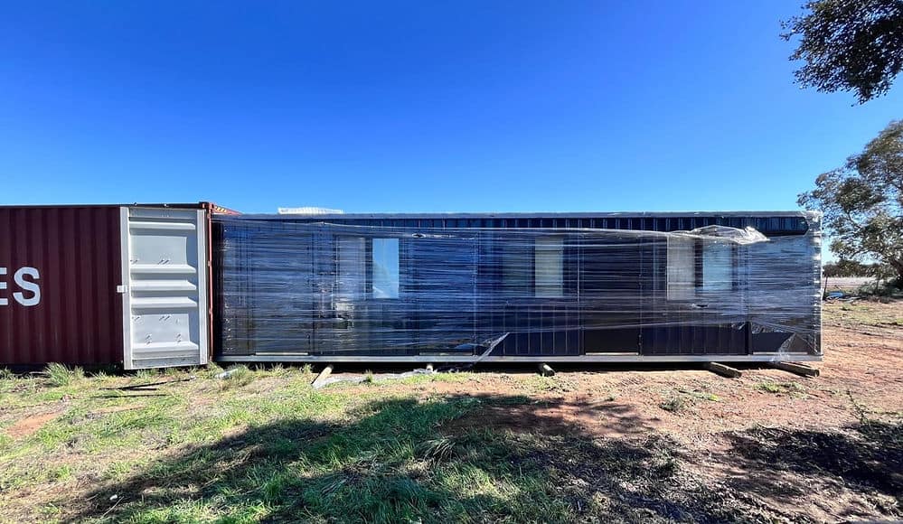 A large, plastic-wrapped shipping container sits on a dirt lot with grass and trees under a blue sky.