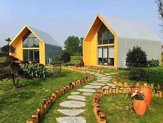 Two small modern houses with curved roofs and large windows, set among green lawns, plants, and a stone path with wood borders.