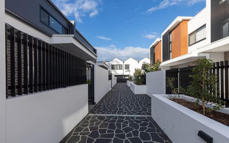 Modern townhouse complex with a paved path, white walls, black railings, and garden beds under a partly cloudy sky.