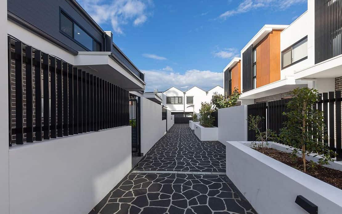 Modern townhouse complex with a paved path, white walls, black railings, and garden beds under a partly cloudy sky.