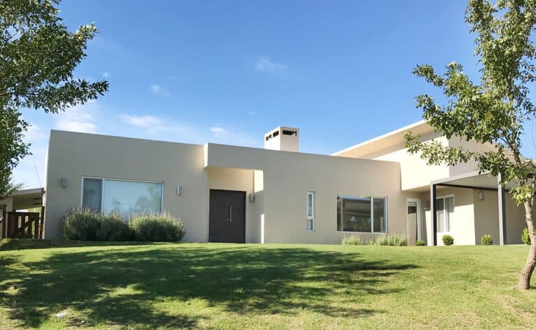 Modern single-story house with flat roof, large windows, light walls, and grassy lawn beneath a clear blue sky.
