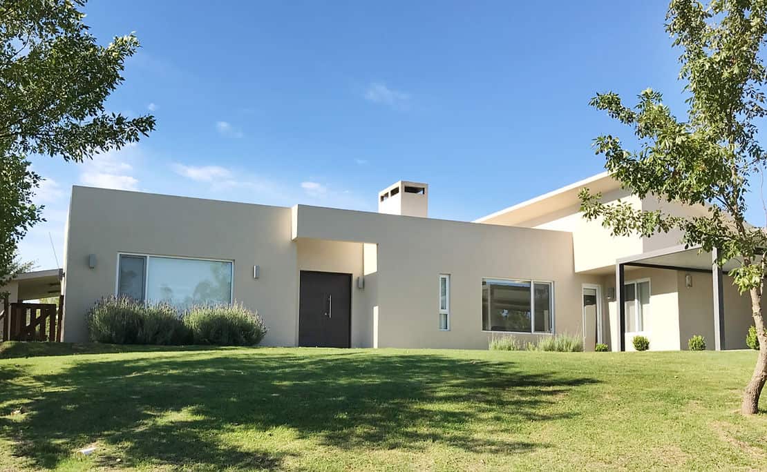 Modern single-story house with flat roof, large windows, light walls, and grassy lawn beneath a clear blue sky.