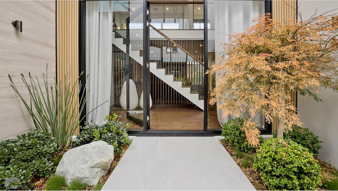 Glass doors reveal a modern interior with a staircase, while plants, an ornamental tree, and rocks frame the entrance.