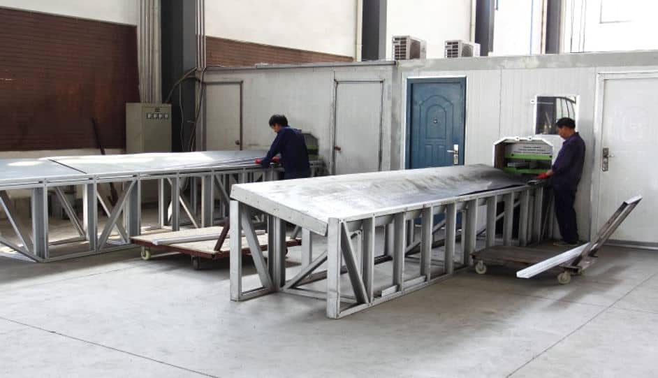 Two workers in an industrial workshop handle large metal sheets and use machinery at separate metal tables.