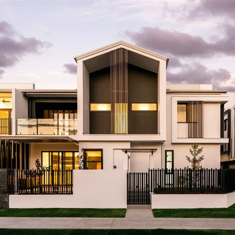 Modern two-story house with large windows, metal accents, small yard, and black fence at sunset.