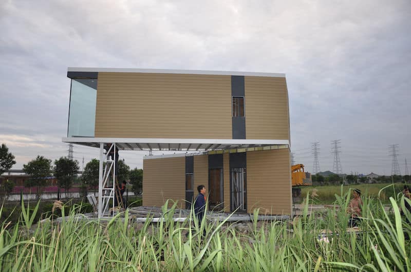Modern two-story house under construction with glass and wood panels, people nearby, grassy field, and power lines in back.