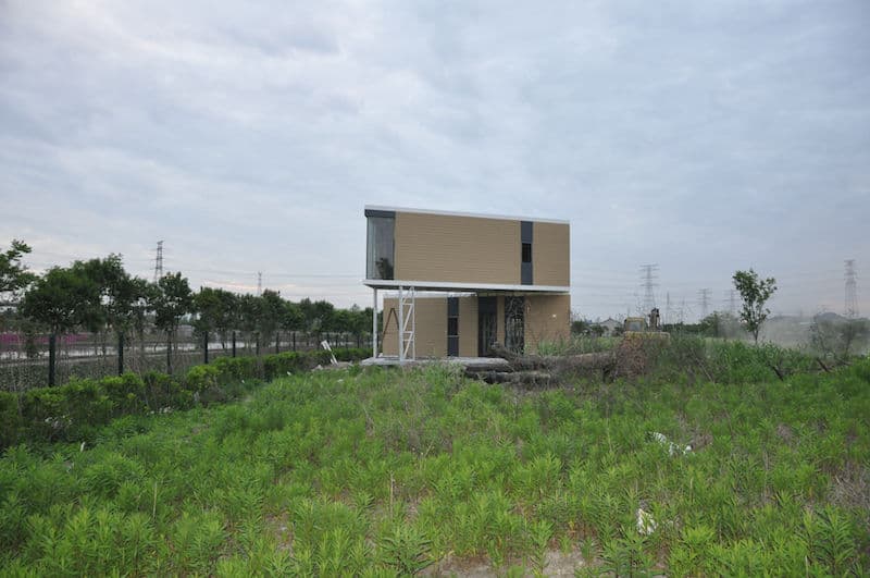 A modern two-story house with large windows sits alone in a grassy field, trees nearby under overcast skies.