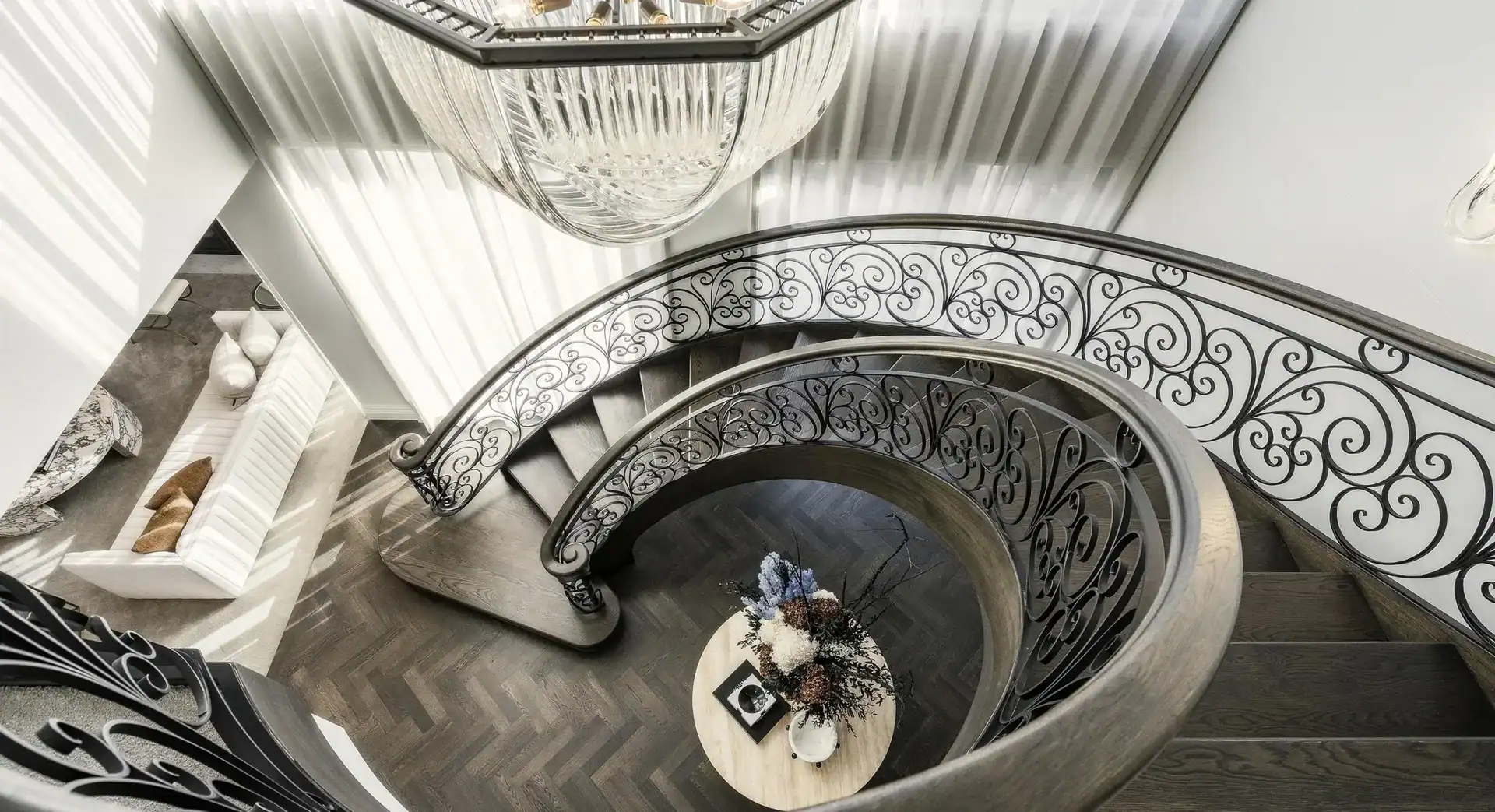 Ornate spiral staircase curves around a table with flowers, viewed from above beneath a large crystal chandelier.