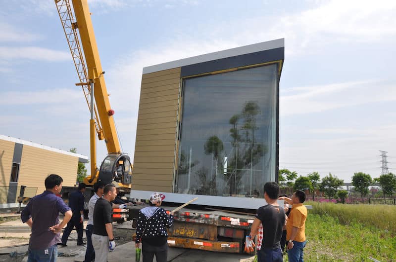 People watch a crane lift a modular building unit onto a flatbed truck at a construction site.