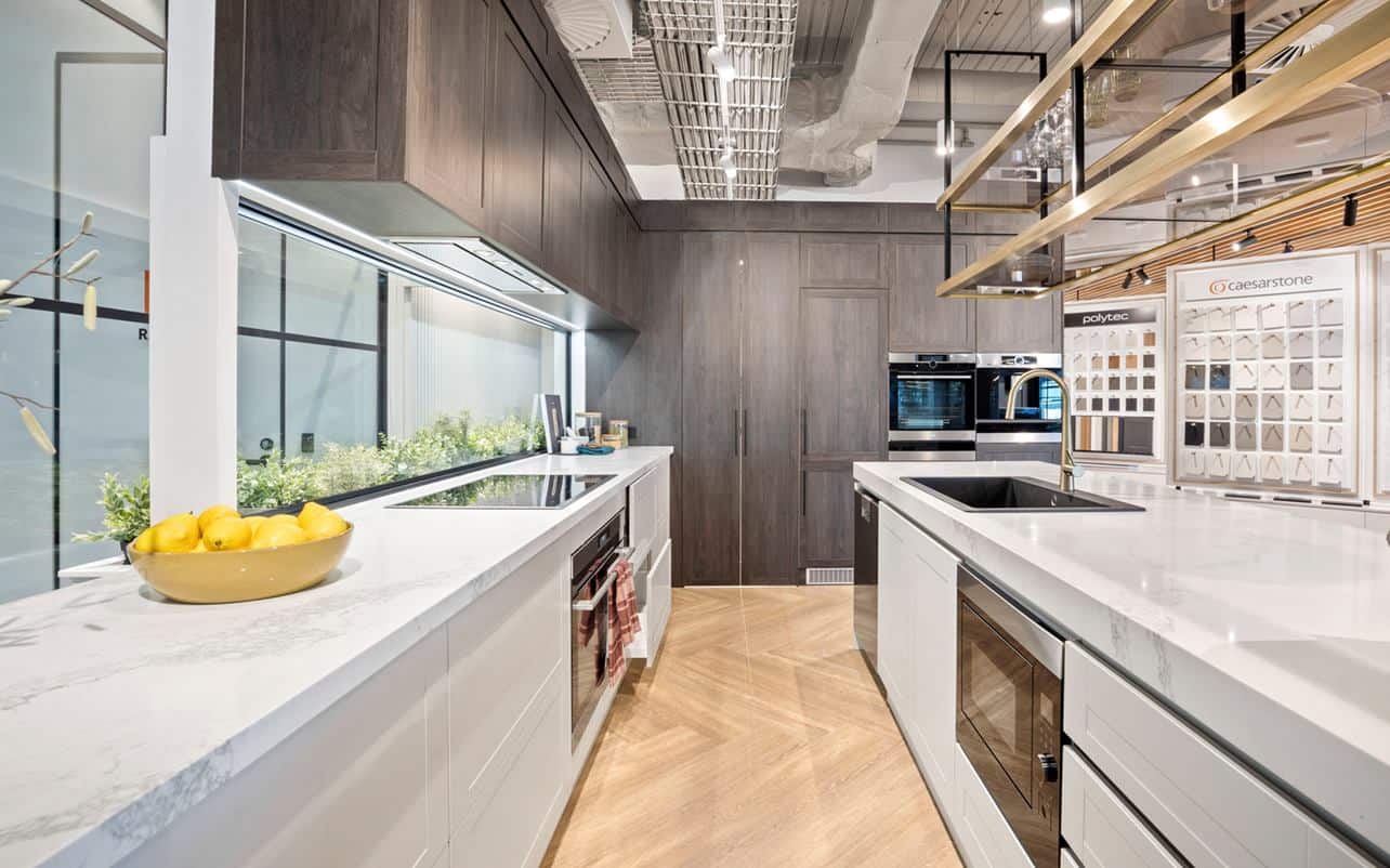 Modern kitchen with white counters, dark wood cabinets, built-in appliances, lemons in a bowl, and window with greenery outside.