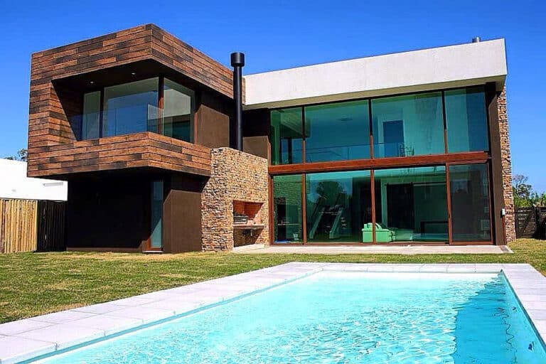 Modern two-story house with glass walls, stone and wood, plus a rectangular pool in the backyard on a clear day.