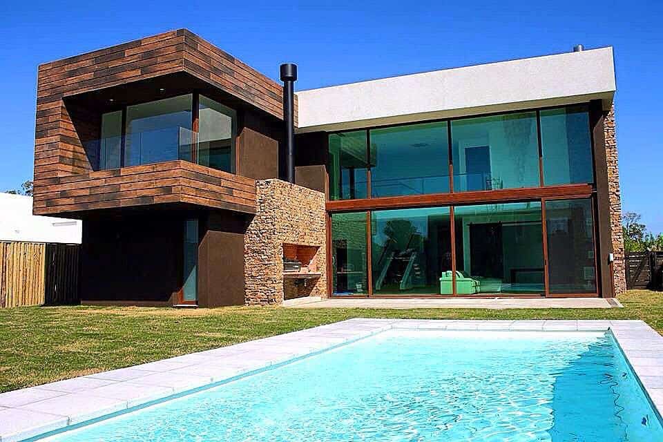 Modern two-story house with glass walls, stone and wood, plus a rectangular pool in the backyard on a clear day.