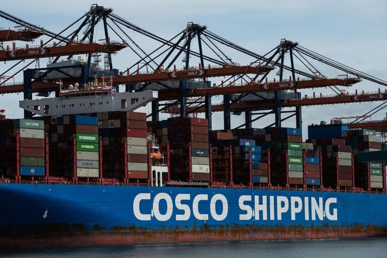 COSCO Shipping container ship docked at port with stacked containers and cranes in the background.