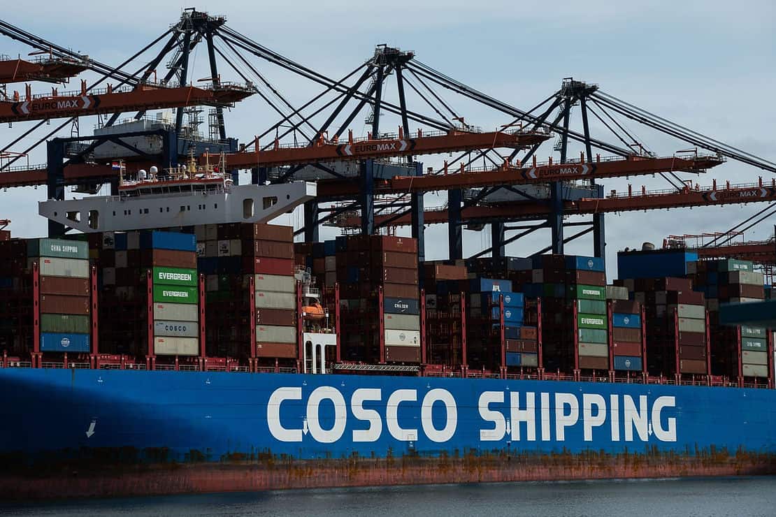 COSCO Shipping container ship docked at port with stacked containers and cranes in the background.