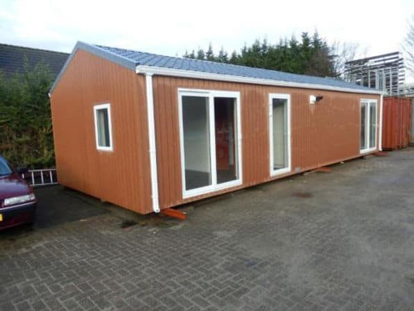 Small brown modular building with sloped roof and glass doors, on pavement by greenery and a nearby car.