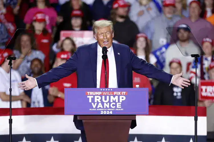 Man in suit speaks at "Trump Vance 2024" podium, audience and American flag visible behind him at a rally.