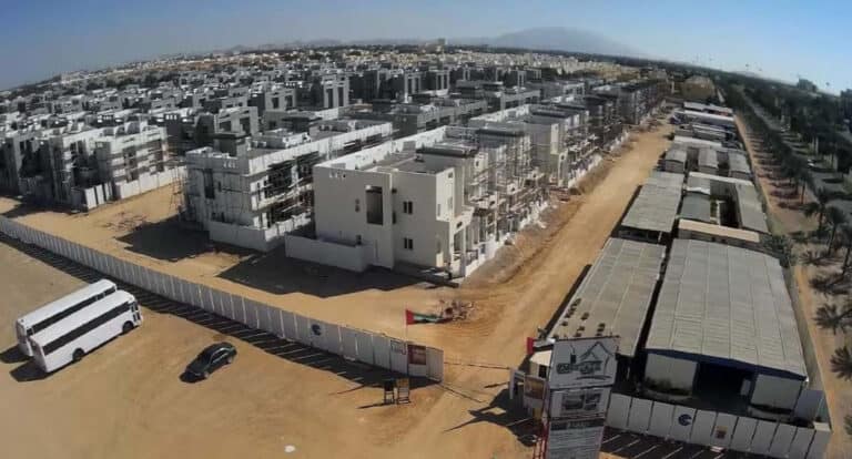 Aerial view of a big housing complex under construction with white buildings, main road, buses, and site fencing.