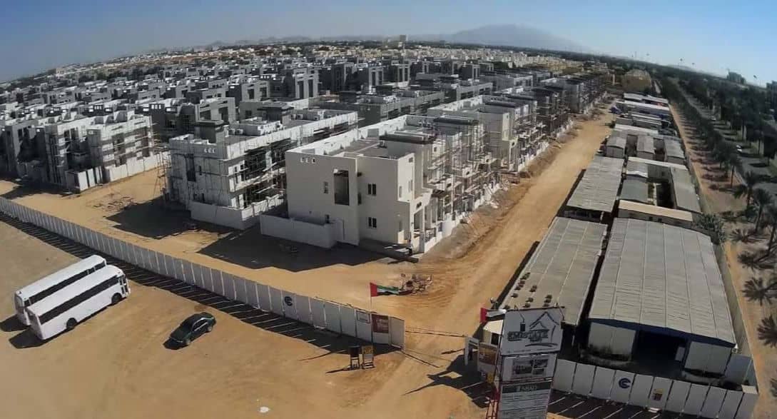 Aerial view of a big housing complex under construction with white buildings, main road, buses, and site fencing.