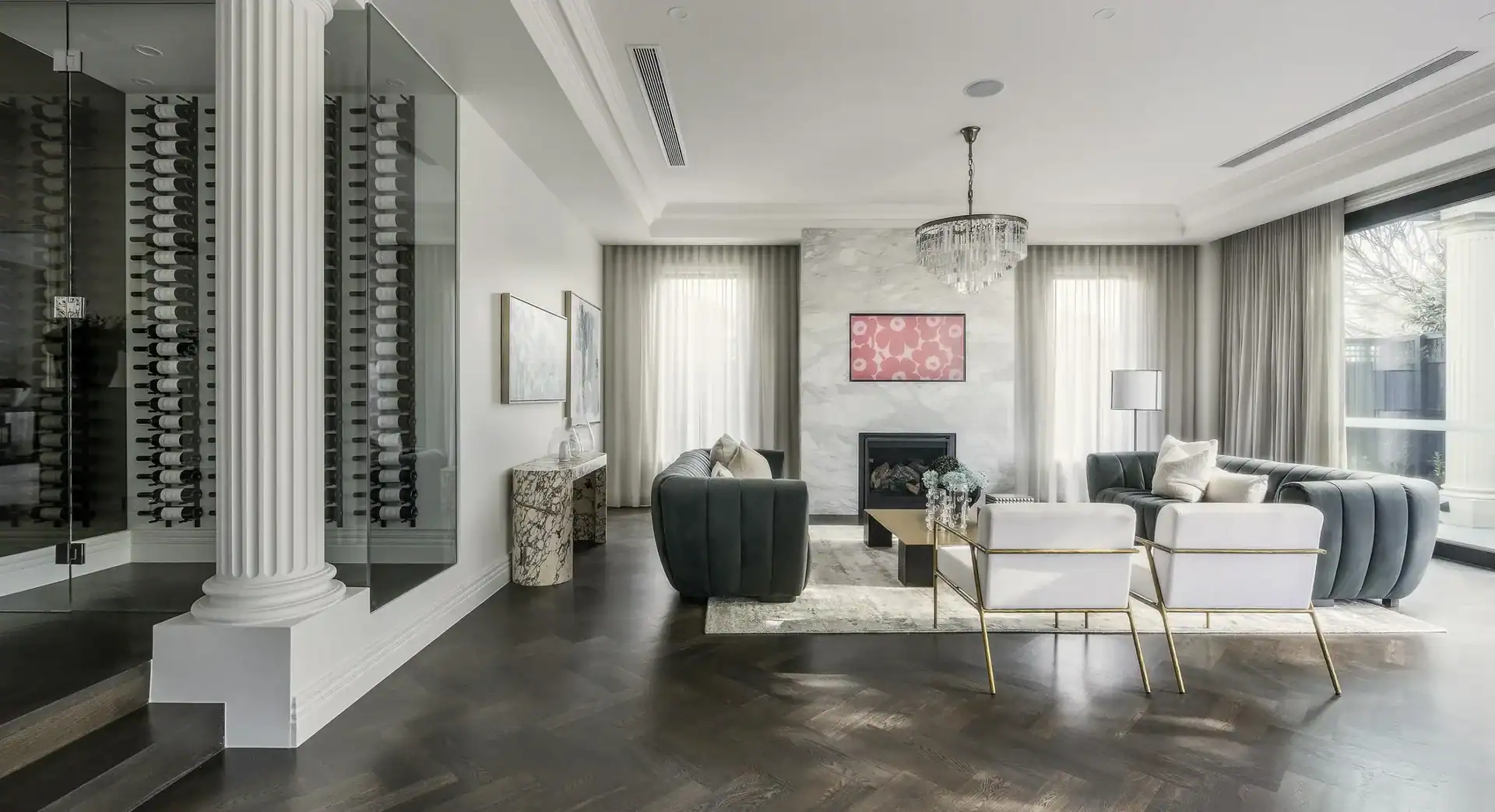 Modern living room with gray sofas, white chairs, glass coffee table, marble fireplace, chandelier, and wine rack.