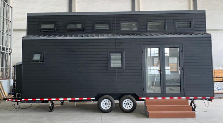 Modern black tiny house on wheels with glass double doors and windows, brown step at entrance, parked indoors.
