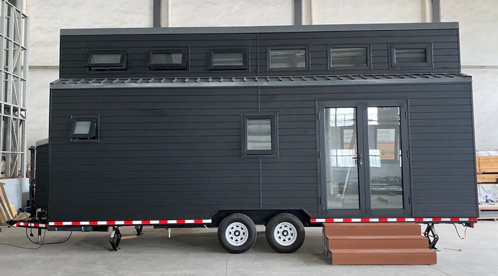 Modern black tiny house on wheels with glass double doors and windows, brown step at entrance, parked indoors.