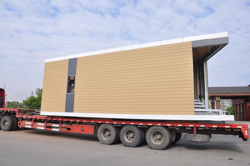 A modular house with open walls and a window is loaded onto a flatbed truck under a cloudy sky.