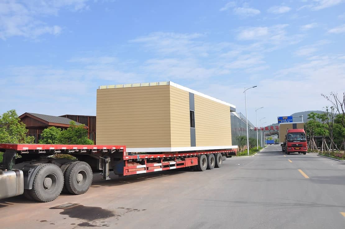 A flatbed truck hauls a rectangular prefab building module along a paved road under a clear sky.