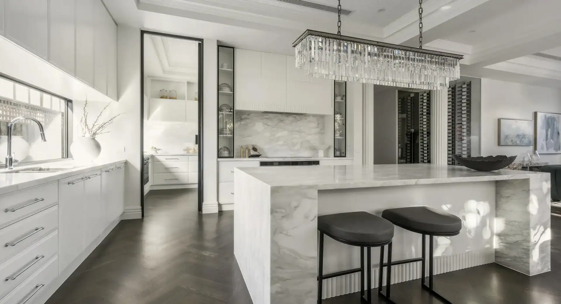 Modern kitchen with white marble island, black stools, white cabinets, stainless appliances, and crystal chandelier.