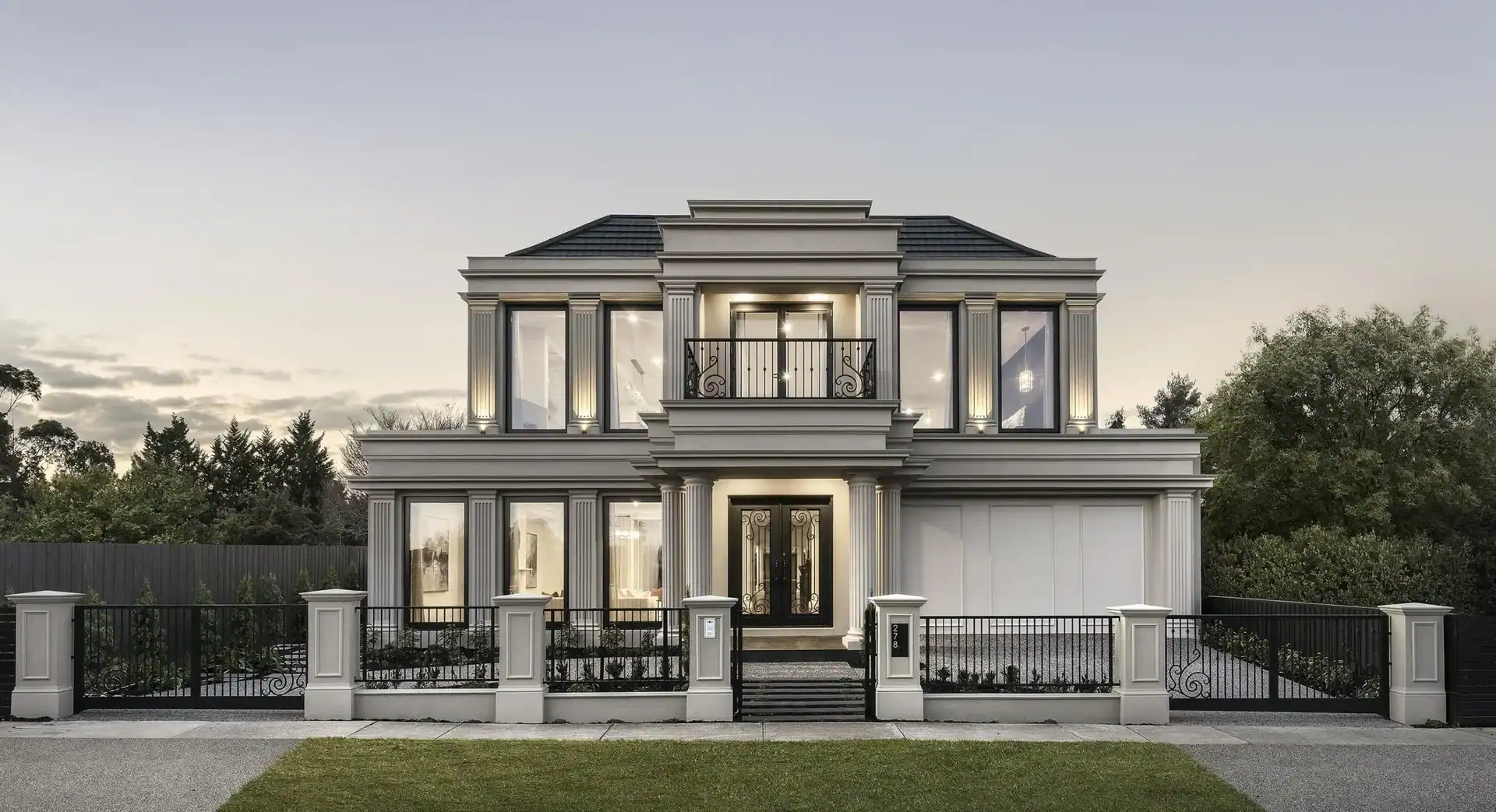 Modern two-story mansion with large windows, beige walls, black roof, and iron fence seen from the lawn at dusk.