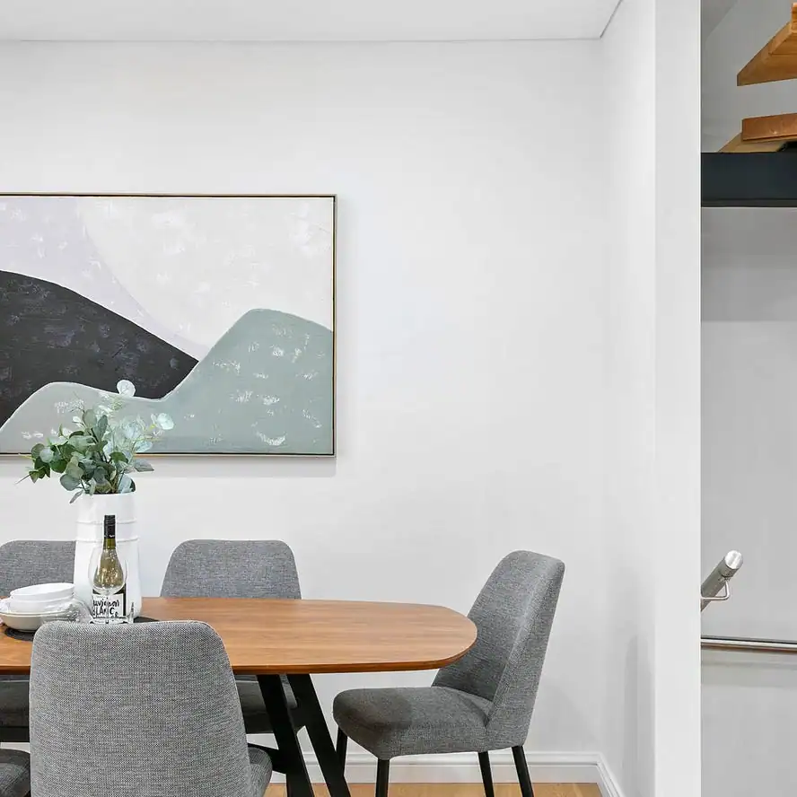 Modern dining area with wooden table, gray chairs, vase of greenery, wine bottle, and abstract art on a white wall.