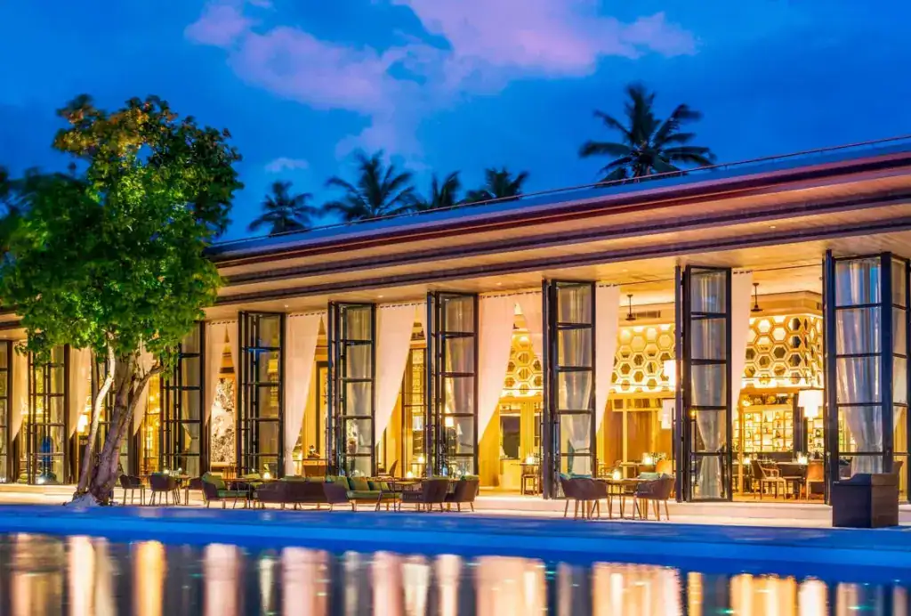 Modern glass building with lit interior, outdoor seating, pool reflections at dusk, and palm trees in the background.