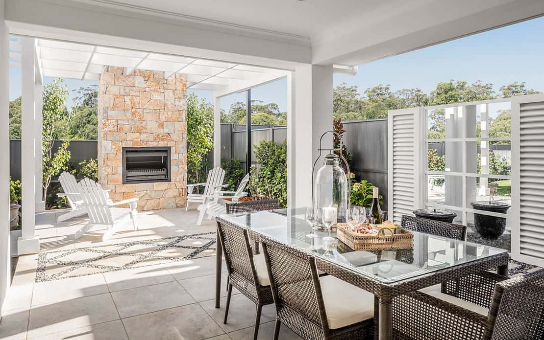 Modern patio with glass table, wicker and lounge chairs, stone fireplace, plants, and privacy fence on a sunny day.