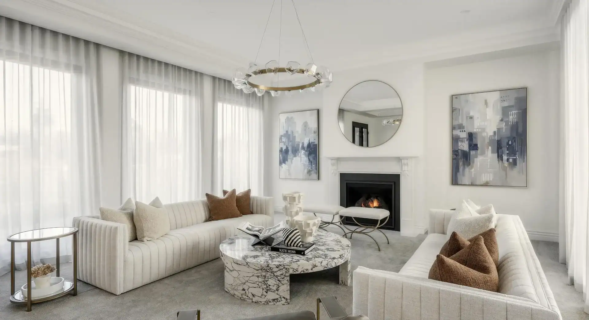 Modern living room with striped sofas, marble coffee table, fireplace, round mirror, abstract art, and large windows.