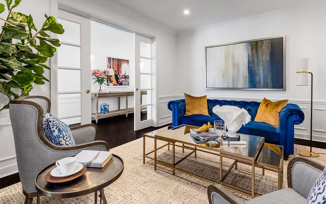 Modern living room with blue velvet sofa, gold accents, glass coffee table, armchair, abstract art, and plant by white doors.