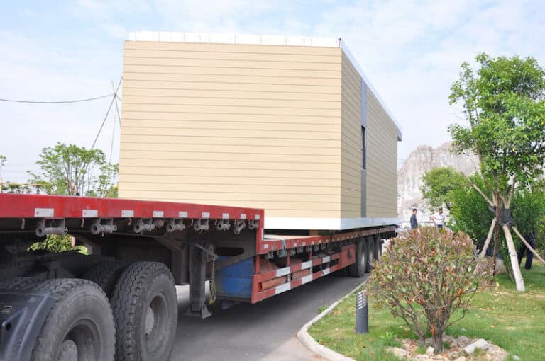 A beige modular building is loaded onto a flatbed truck parked on a road lined with trees and plants.