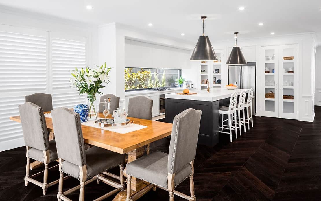Modern kitchen with wooden dining table, six chairs, island with stools, pendant lights, and white cabinets with steel appliances.
