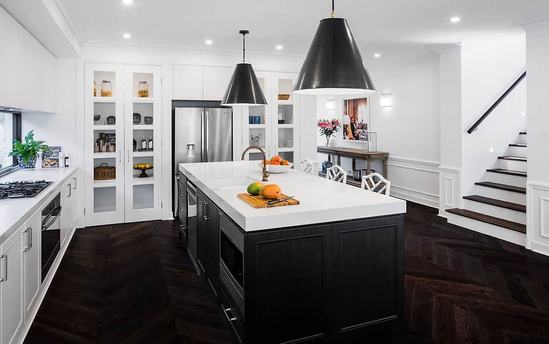 Modern kitchen with island, black pendants, white cabinets, stainless appliances, dark wood floors, and a staircase on the right.