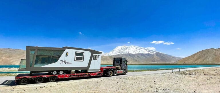 A truck transports a modern mobile home on a flatbed by a lake, with mountains and a snow-capped peak in the background.