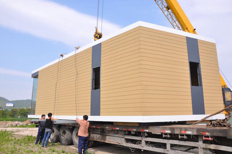 A crane lifts a modular building onto a flatbed truck as three workers guide the placement.