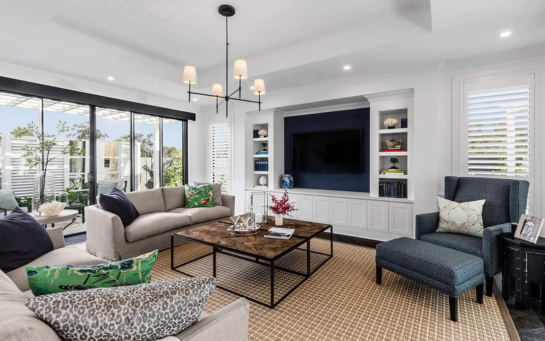Modern living room with beige sofas, blue armchair, rug, coffee table, TV, shelves, and large windows overlooking a garden.