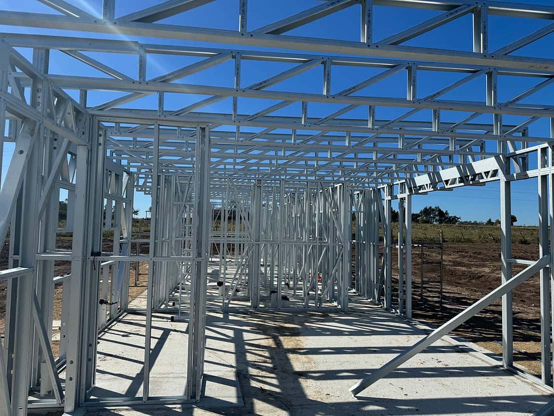 A metal frame building under construction on a concrete base, with open land and a clear sky in the background.