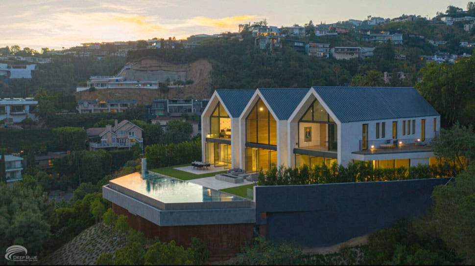 Modern hillside home with glass windows, infinity pool, scenic views, trees, and neighbors at sunset.