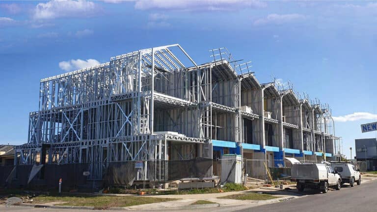 Multi-story buildings under construction with steel frames, scaffolding, and vehicles parked nearby under a blue sky.